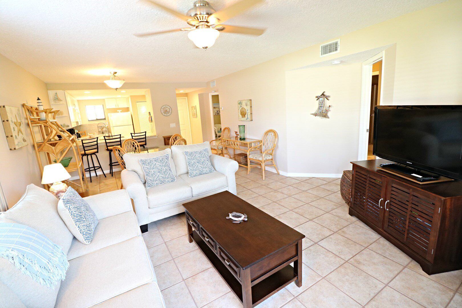 A spacious living room with a sofa, coffee table, and TV stand. There is a dining area with chairs and a table in the background.