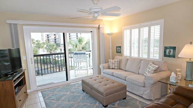 A living room with a sofa, ottoman, television, and a balcony with a view of palm trees. - Ocean Village Club H31 (St. Augustine)
