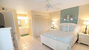 A bedroom with a bed, nightstands, a ceiling fan, a dresser, a closet, and a bathroom with a sink and mirror. - Ocean Village Club H31 (St. Augustine)