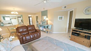 A living room with a brown leather sofa, a beige ottoman, a wooden TV stand with a flat-screen TV, and a dining area with wicker chairs. - Ocean Village Club H31 (St. Augustine)