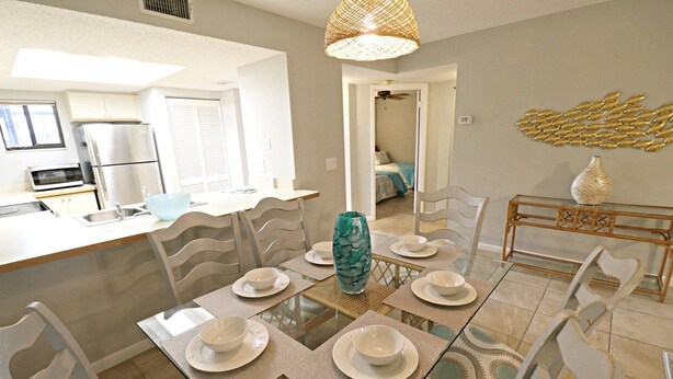 A dining area with a glass table set for six, surrounded by white chairs. A large vase is placed in the center. - Ocean Village Club Condo J16 (St. Augustine)