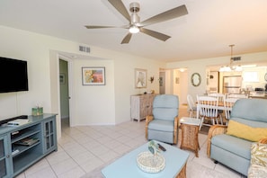 A living room with a TV, a ceiling fan, a dining area with chairs and a table, and a kitchen in the background.