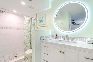 Un baño con inodoro, ducha con cortina estampada, lavamanos y espejo, y un ventilador de techo.