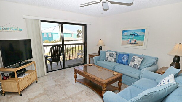 A living room with a TV, sofa, coffee table, and a balcony with chairs. - Ocean Village Club Condo L24 (St. Augustine)