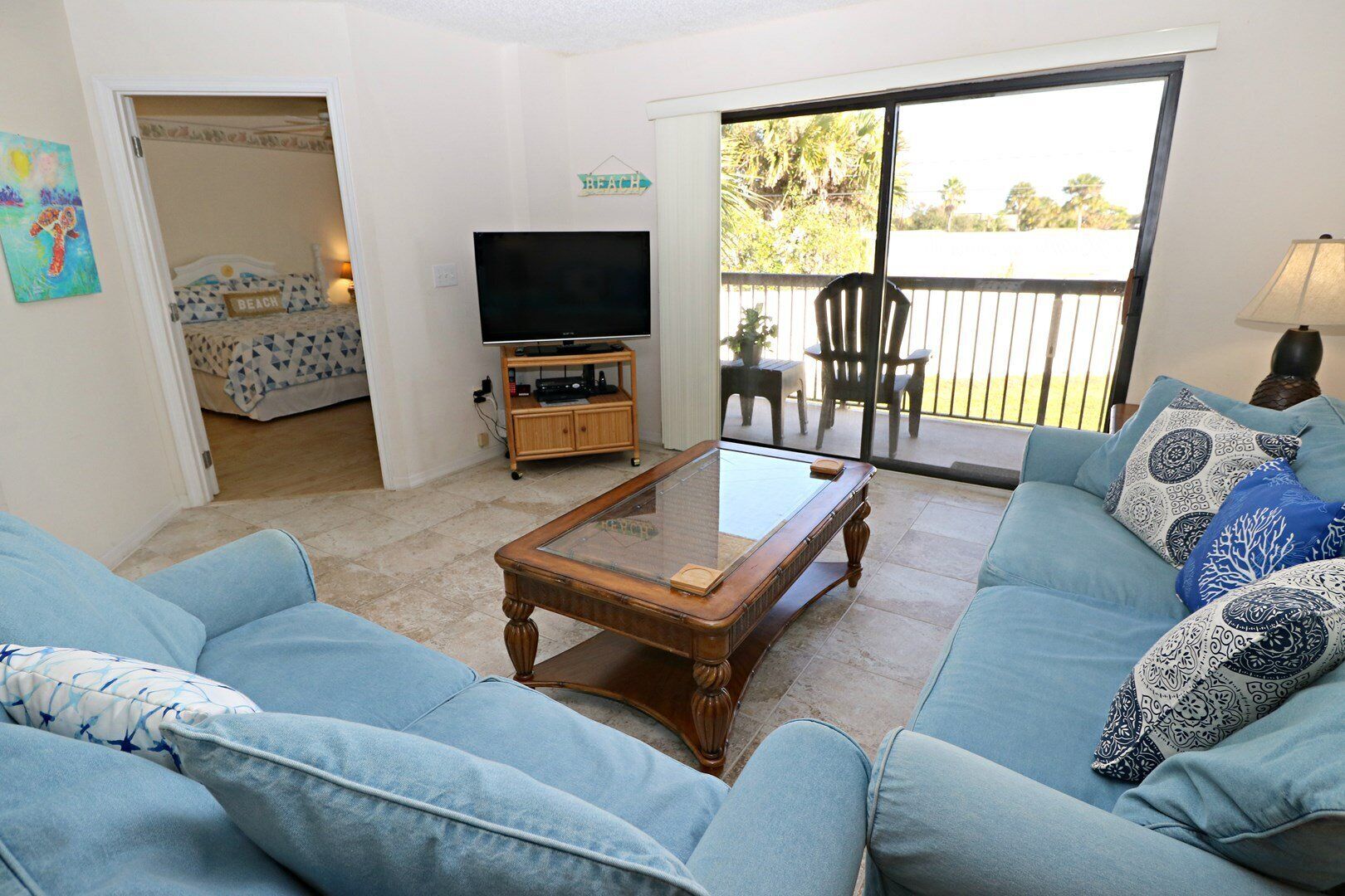 A living room with a glass-top coffee table, a sofa, a television, and a balcony with outdoor furniture.