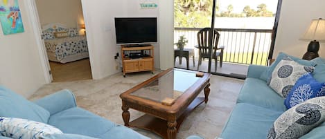 A living room with a glass-top coffee table, a sofa, a television, and a balcony with outdoor furniture.