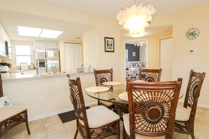 A dining area with a glass table and wooden chairs, a kitchen in the background, and a chandelier.