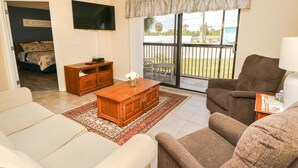 A living room with a sofa, armchair, wooden coffee table, and a TV mounted on the wall. There is a sliding glass door leading to a balcony with outdoor furniture. - Ocean Village Club Condo L26 (St. Augustine)