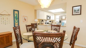 A dining area with a glass table and wicker chairs, a kitchen in the background, and a chandelier. - Ocean Village Club Condo L26 (St. Augustine)