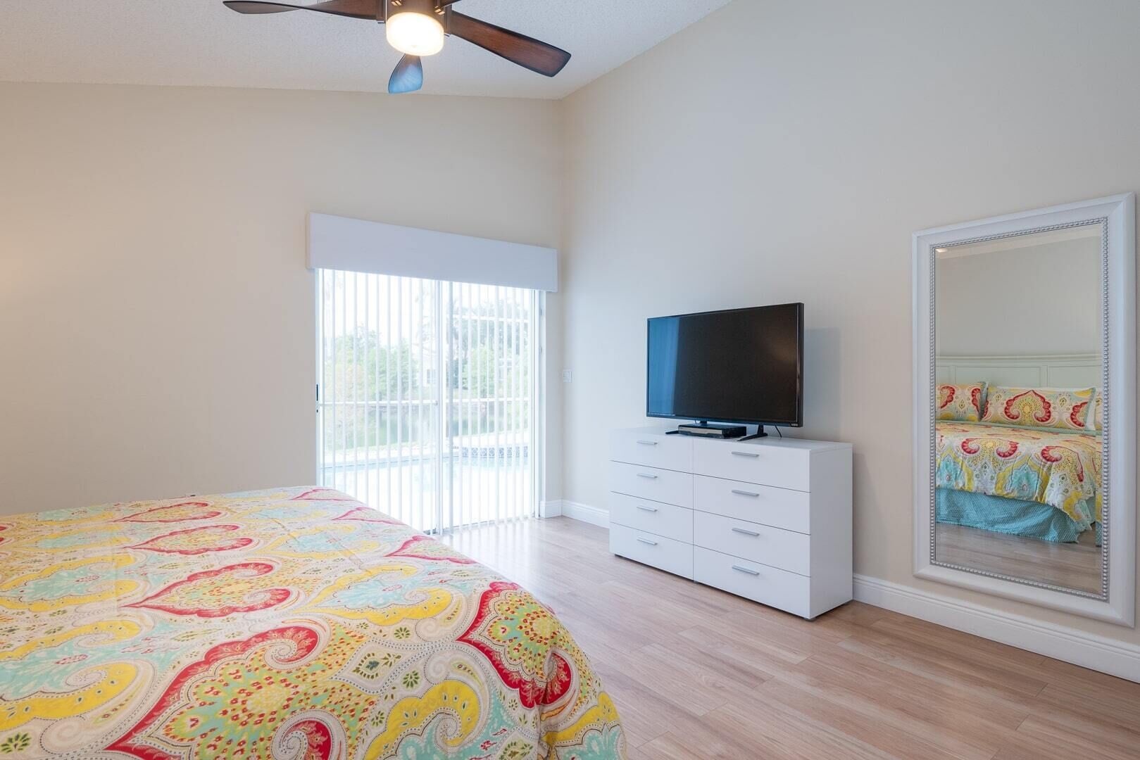 A bedroom with a bed, a television on a white dresser, a large mirror, and a ceiling fan.