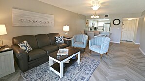 A modern living room with a brown leather sofa, two armchairs, a coffee table, and a patterned rug. A kitchen is visible in the background with a dining area. - Ocean Village Club Condo O32 (St. Augustine)