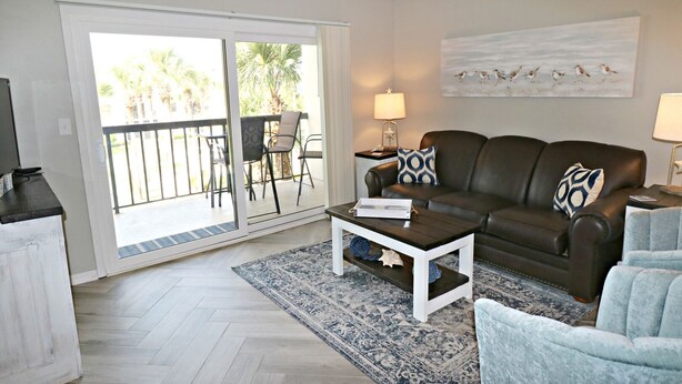 A living room with a brown leather sofa, a white coffee table, and a blue armchair. There is a sliding glass door leading to an outdoor area with a patio table and chairs. - Ocean Village Club Condo O32 (St. Augustine)