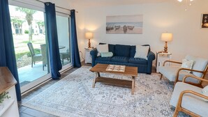 A living room with a blue sofa, a wooden coffee table, and a patterned rug. There are two lamps on side tables, a painting on the wall, and a view of the outdoors through a large window with blue curtains. - Ocean Village Club Condo P18 (St. Augustine)