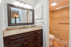 Une salle de bain dotée d’un lavabo avec un plan de travail en granit, d’un meuble-lavabo en bois foncé, d’un grand miroir avec éclairage intégré, de toilettes et d’une cabine de douche en verre.
