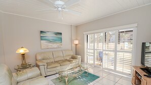 Ein Wohnzimmer mit einem beigefarbenen Sofa, einem Glastisch, einer Lampe, einem Strandbild und einem Fernseher.