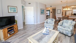 A living room with a TV, two patterned armchairs, a coffee table, and a dining area with a table and chairs.
