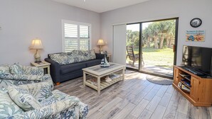 A living room with a sofa, coffee table, and TV stand. There is a sliding glass door leading to a garden.