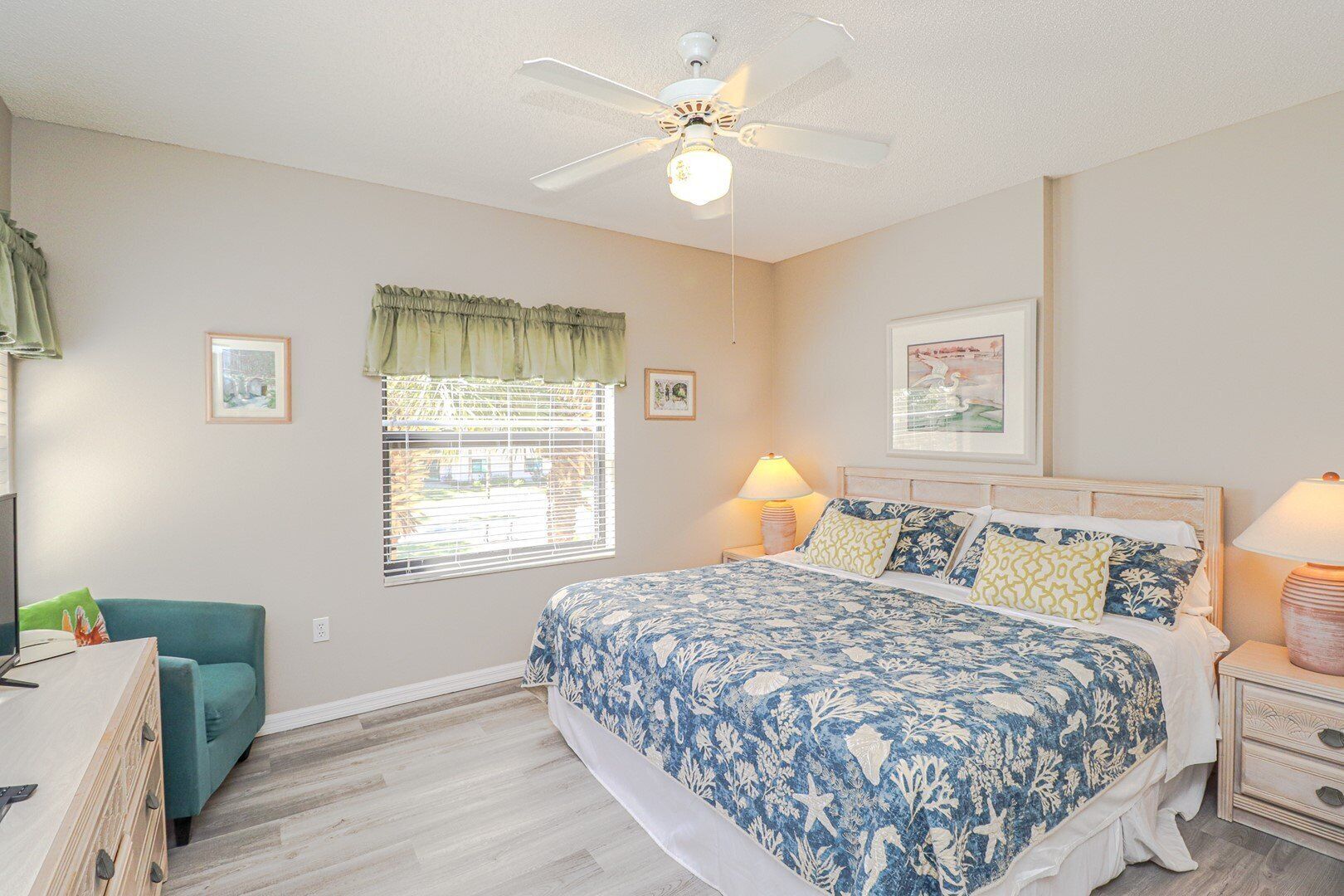 A bedroom with a bed, a desk, a chair, a ceiling fan, a window with blinds, and a lamp.