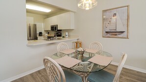 Un coin repas avec une table en verre, des chaises blanches, un lustre, une cuisine en arrière-plan et un tableau encadré au mur.
