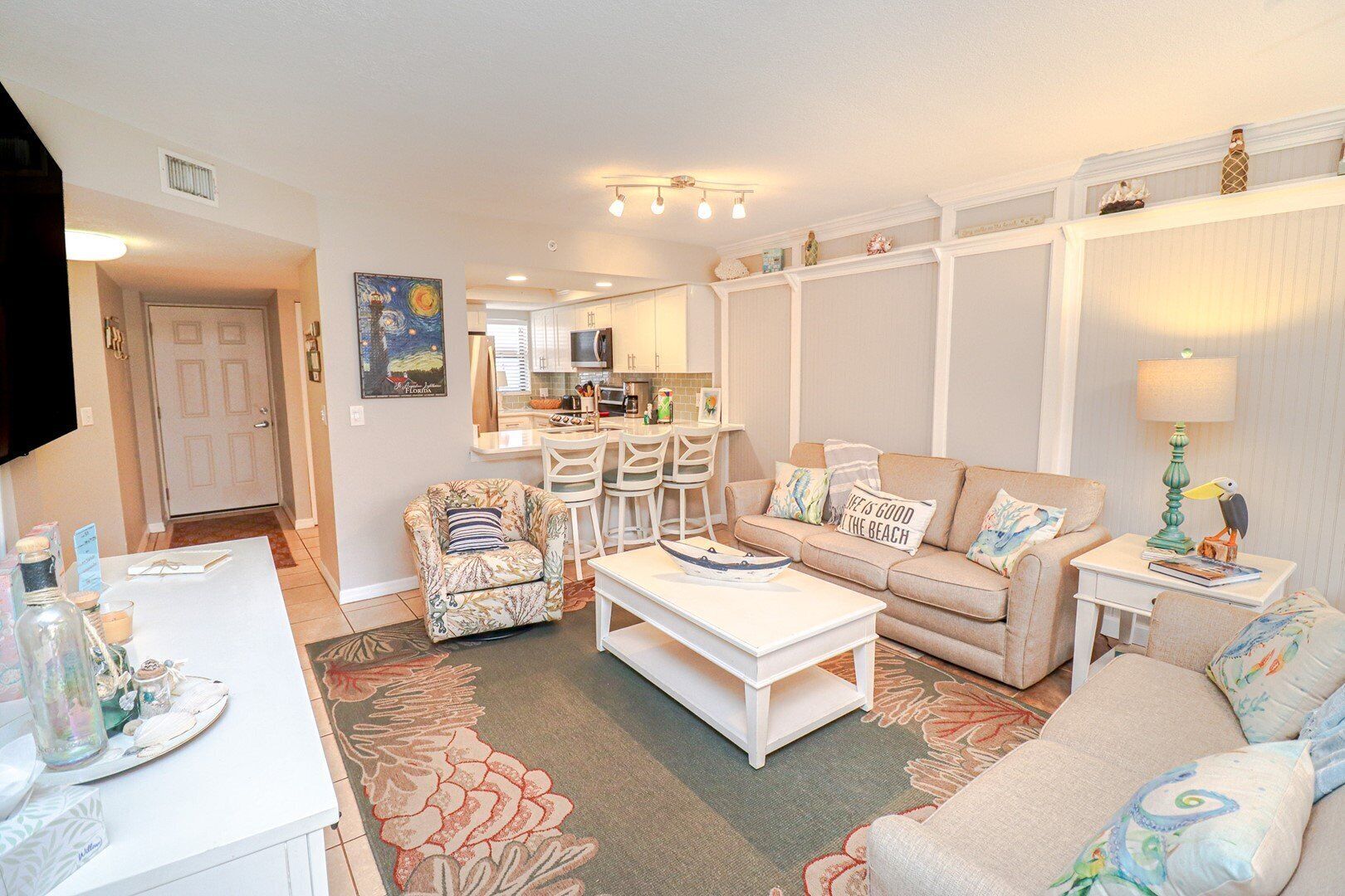 A living room with a sofa, two armchairs, a coffee table, and a dining table. There is a kitchen in the background with a microwave and a sink.