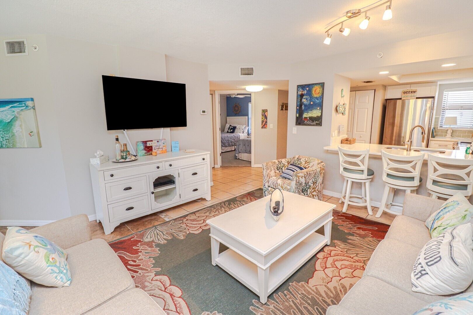 A living room with a flat-screen TV, a white entertainment unit, a coffee table, and a sofa. There is a kitchen area with a bar and chairs.