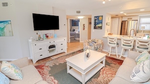 A living room with a flat-screen TV, a white entertainment unit, a coffee table, and a sofa. There is a kitchen area with a bar and chairs.