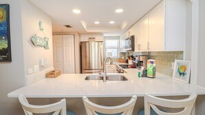 A modern kitchen with white cabinetry, a stainless steel refrigerator, and a marble countertop. There are three white chairs with blue cushions.