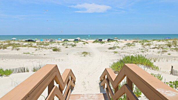 A beach pathway leading to the ocean with beach umbrellas and cars in the background.