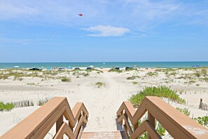 A beach pathway leading to the ocean with beach umbrellas and cars in the background.