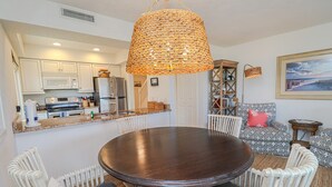 A dining area with a round table surrounded by white chairs, a kitchen in the background, and a large woven light fixture above.