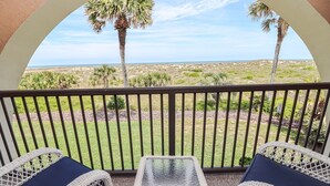 A balcony with wicker furniture and a glass table, overlooking a beach with palm trees and a clear sky.