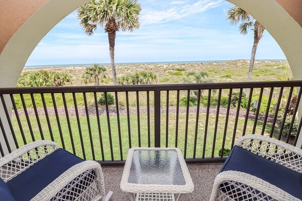 A balcony with wicker furniture and a glass table, overlooking a beach with palm trees and a clear sky.