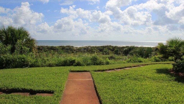 Ein Weg führt zu einem Strand mit freiem Blick auf das Meer und einer Rasenfläche mit Palmen.