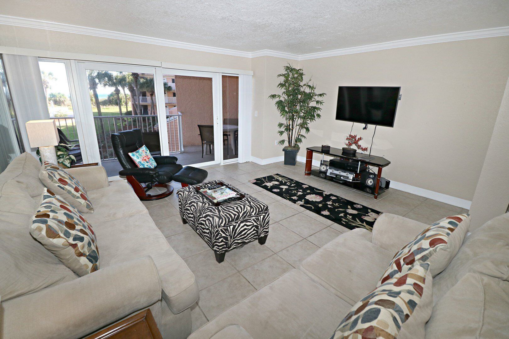A living room with a beige sofa, a black leather chair, a zebra-patterned ottoman, a TV on a stand, and a potted plant.