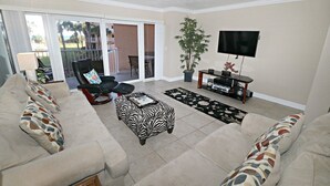 Ein Wohnzimmer mit einem beigen Sofa, einem schwarzen Ledersessel, einem Ottomanen mit Zebra-Muster, einem Fernseher auf einem Schrank und einer Zimmerpflanze.
