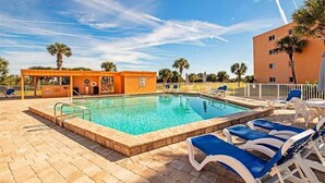 Ein Schwimmbecken mit Liegestühlen, einer überdachten Liegefläche und einem Gebäude im Hintergrund mit Palmen.