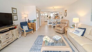 Ein Wohnzimmer mit Fernseher, Sofas, einem Couchtisch mit Blumen und einem Essbereich im Hintergrund.