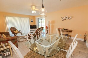 Un salon comprenant une table à manger avec un plateau en verre, entourée de chaises en osier blanc, un canapé et un téléviseur.