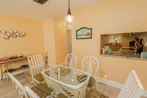 A dining area with a glass table and white chairs, a kitchen visible through an open doorway, and a wall-mounted sign indicating a beach retreat.
