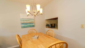 Un comedor con una mesa de madera y cuatro sillas, una araña, un cuadro de paisaje marino y un estante pequeño con adornos.