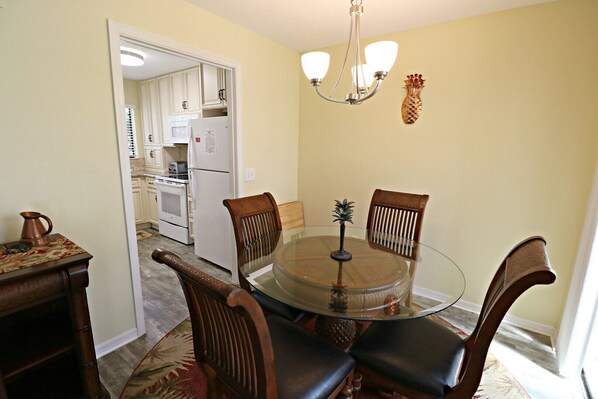 A dining area with a glass table and wooden chairs, a kitchen in the background, and a pineapple wall decoration. - Gorgeous Renovated Pet Friendly Townhome-SABTR 810 (St. Augustine)