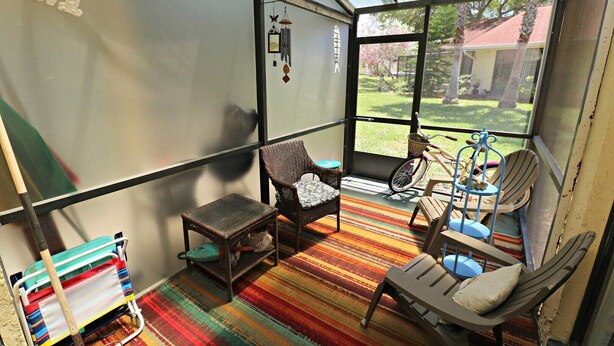 A sunlit room with wicker furniture, a colorful striped rug, and a view of a garden through large windows.