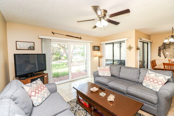 A living room with a grey sofa, a television, a ceiling fan, and a view of a garden through the sliding glass door.