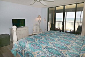 A bedroom with a large bed, a television, a dresser, and a view of the beach through a sliding glass door. - Sand Dollar 2-302 Private Balcony, Direct Oceanfront, Beach Views (St. Augustine)