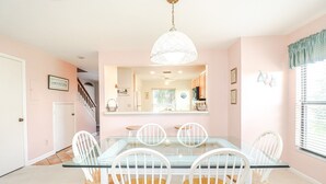 Zona pranzo con tavolo in vetro e sedie bianche, una lampada a sospensione e cucina visibile attraverso un'apertura.