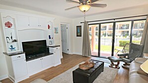 A modern living room with a flat-screen TV, white cabinetry, a ceiling fan, and a view of a residential area through large glass doors. - Updated, Classy Ground Floor Condo-Sea Place 12119 (St. Augustine)