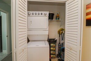 A laundry room with stacked washers and dryers, a shelf with cleaning supplies, and a mop and broom.
