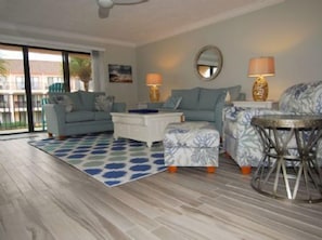 A living room with a sofa, ottoman, coffee table, and a small table with a lamp. There is a large window with a view of buildings outside. - Luxury at the Beach, Totally Remodeled with Ocean Views Sea Place 12214! (St. Augustine)