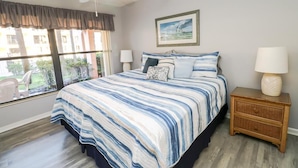 A bedroom with a large bed, two bedside tables, a ceiling fan, and a window with a view of greenery.