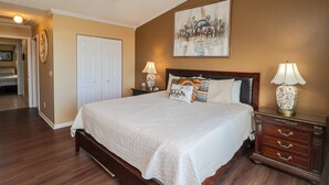 Une chambre à coucher avec un grand lit, deux tables de chevet avec des lampes, un ventilateur de plafond, une porte et un tableau au mur.